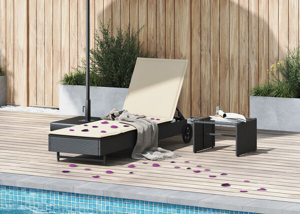 Rattanliege und Tisch am Pool mit weissen Kissen und verstreuten violetten Bl&uuml;tenbl&auml;ttern.