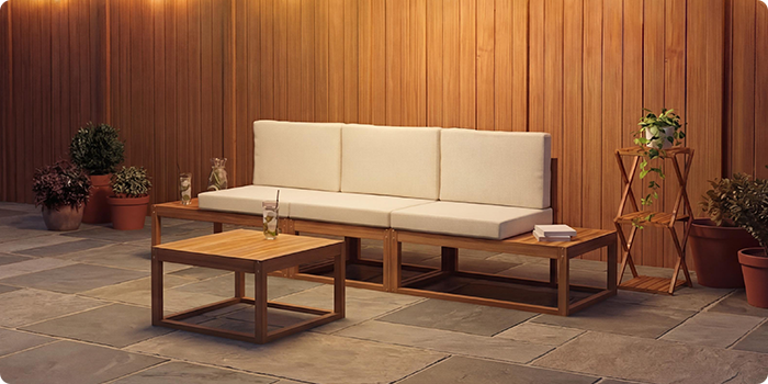 Dreiteiliges modulares Outdoor-Sofa mit cremefarbenen Kissen und passendem niedrigen Couchtisch auf einer Steinterrasse vor einer warmen Holzwand